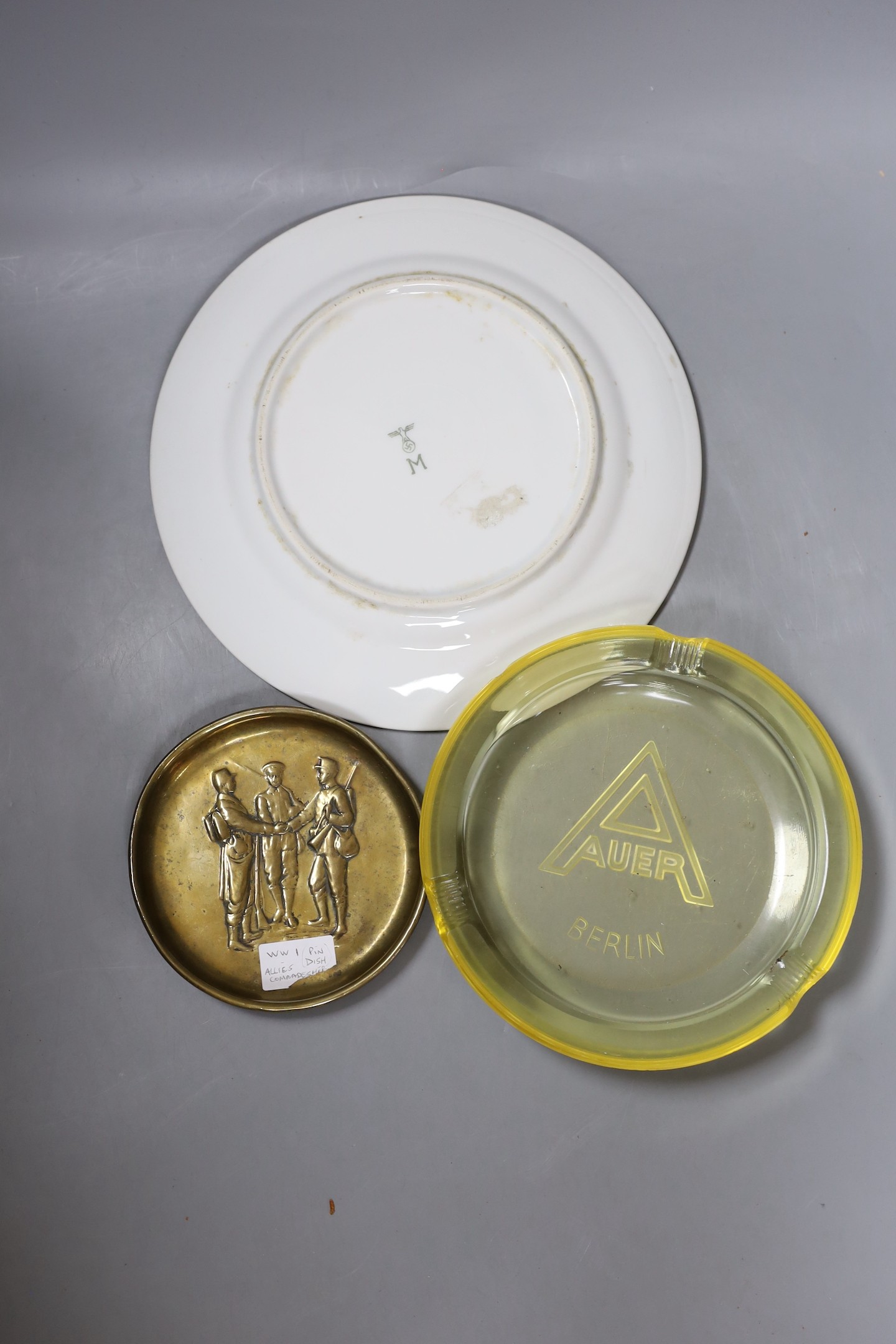A WWII German Mess Hall porcelain plate, an Auer Berlin yellow glass ash tray and a WWI trench art ash tray, largest 24cm diameter